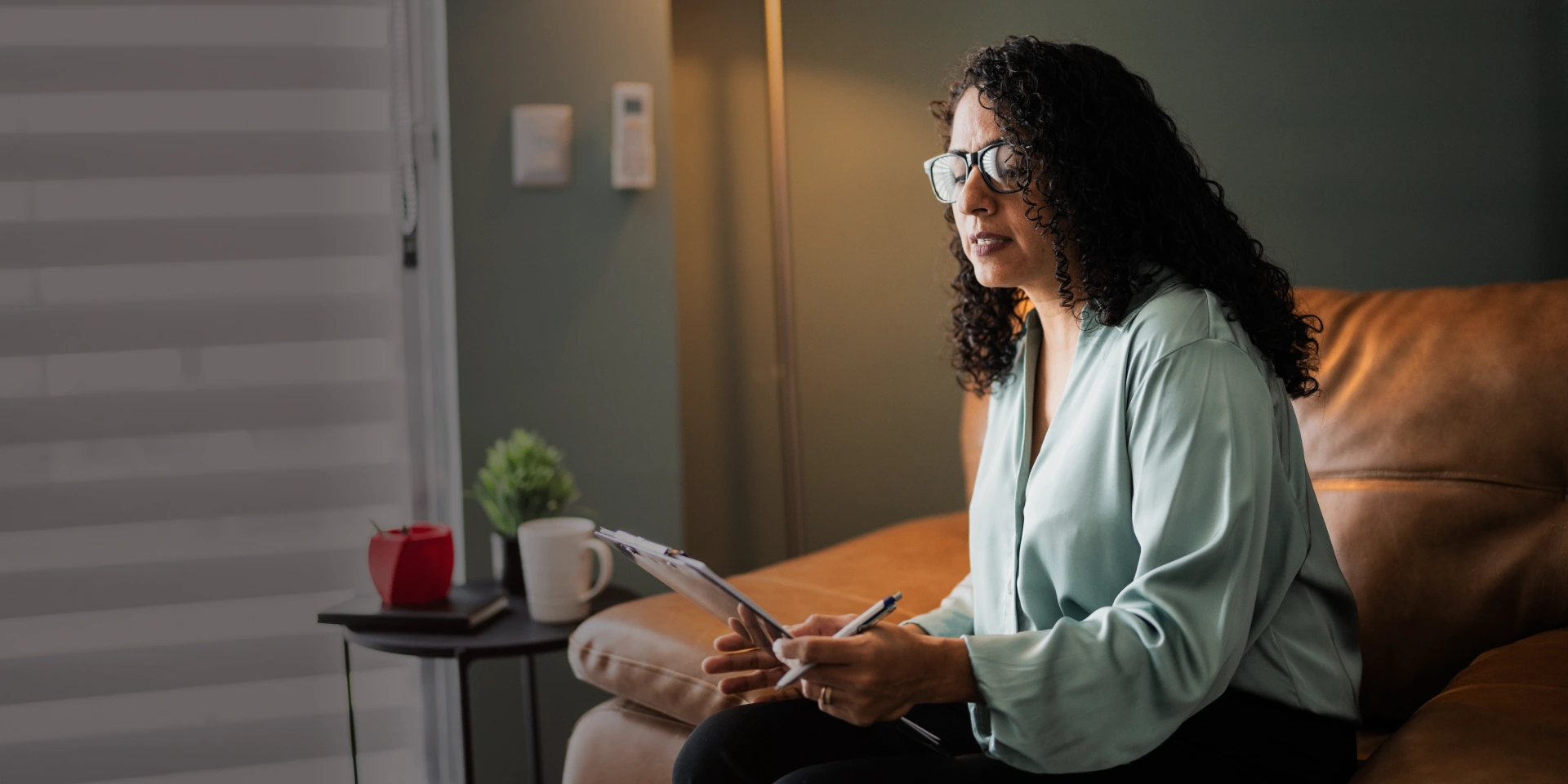 A woman with glasses sits on a couch holding a clipboard and pen, appearing to listen and take notes. A side table with a mug and plant is next to her.