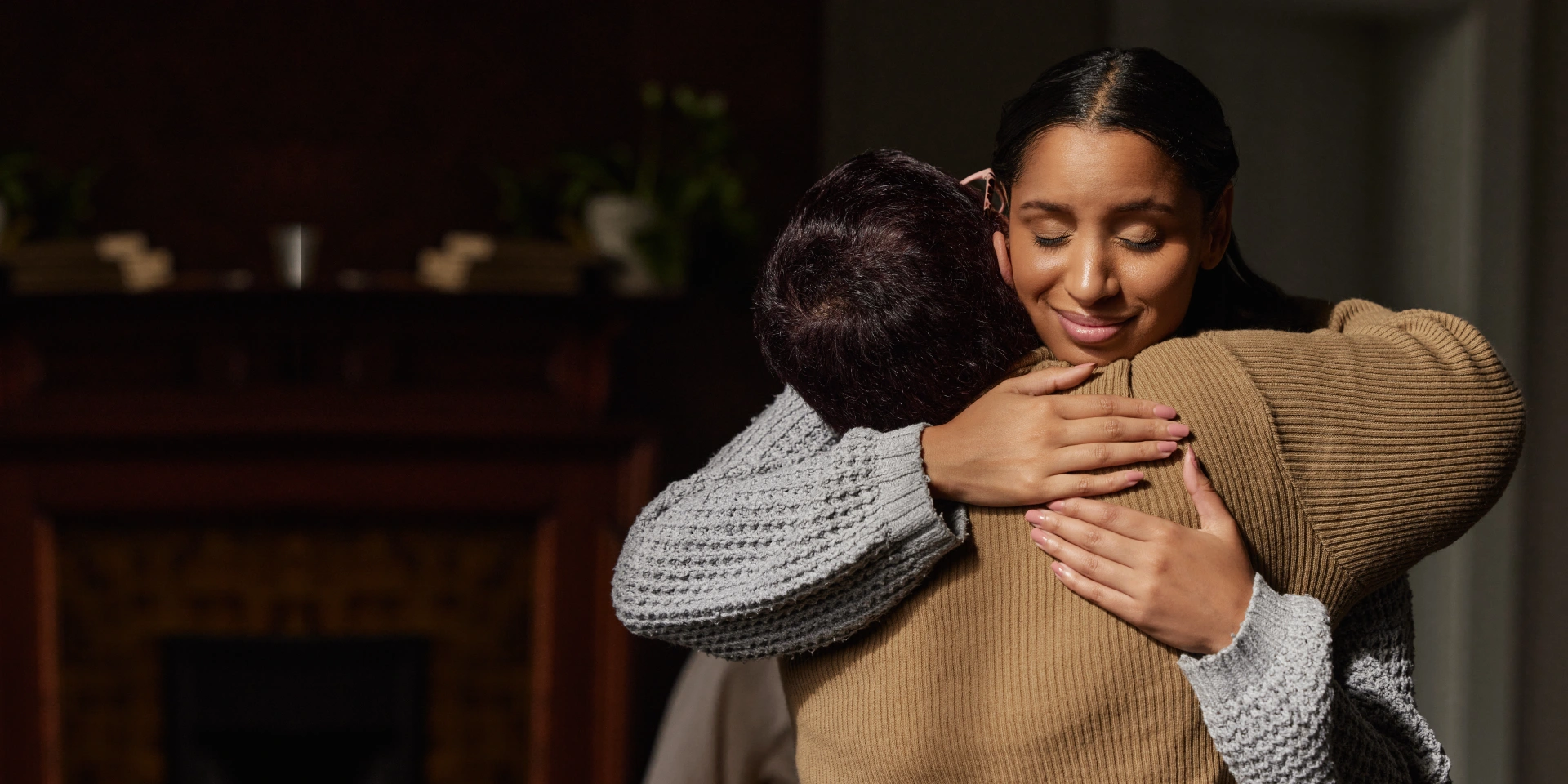 Two people are embracing each other indoors, with one person's face visible and eyes closed, wearing a light sweater in a warmly lit room.