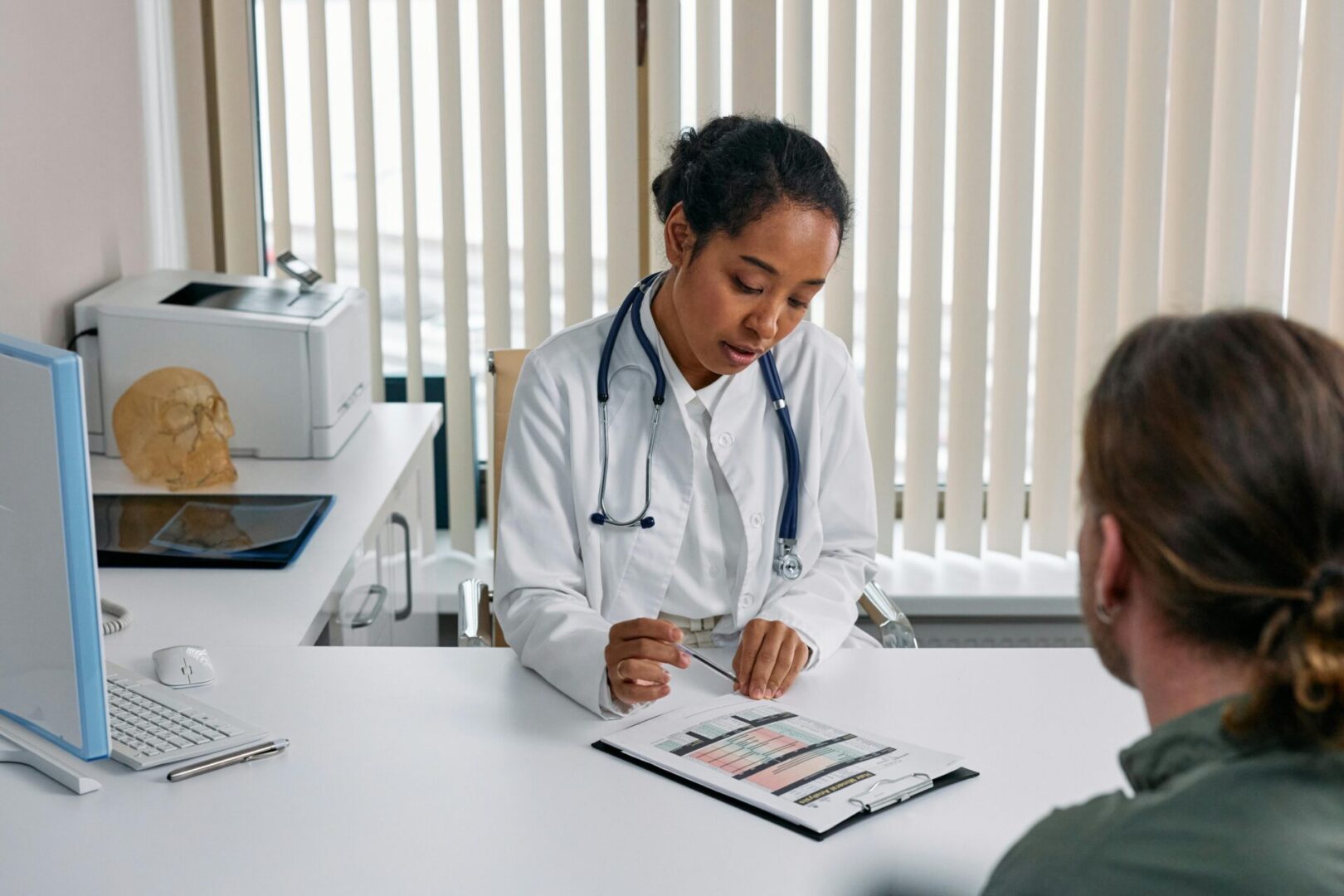 A doctor in a white coat reviews a chart and discusses information with a patient sitting across a desk in a medical office.