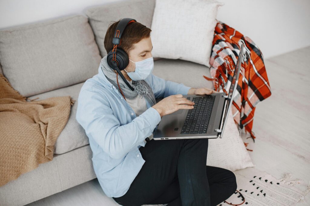 A man wearing a face mask and headphones sits on the floor by a couch, using a laptop.