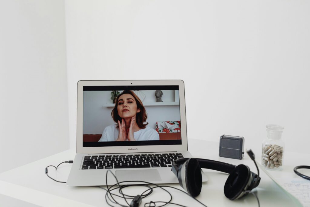 A laptop displays a woman speaking during a video call. Headphones, a small speaker, and a jar of capsules are on the white desk.