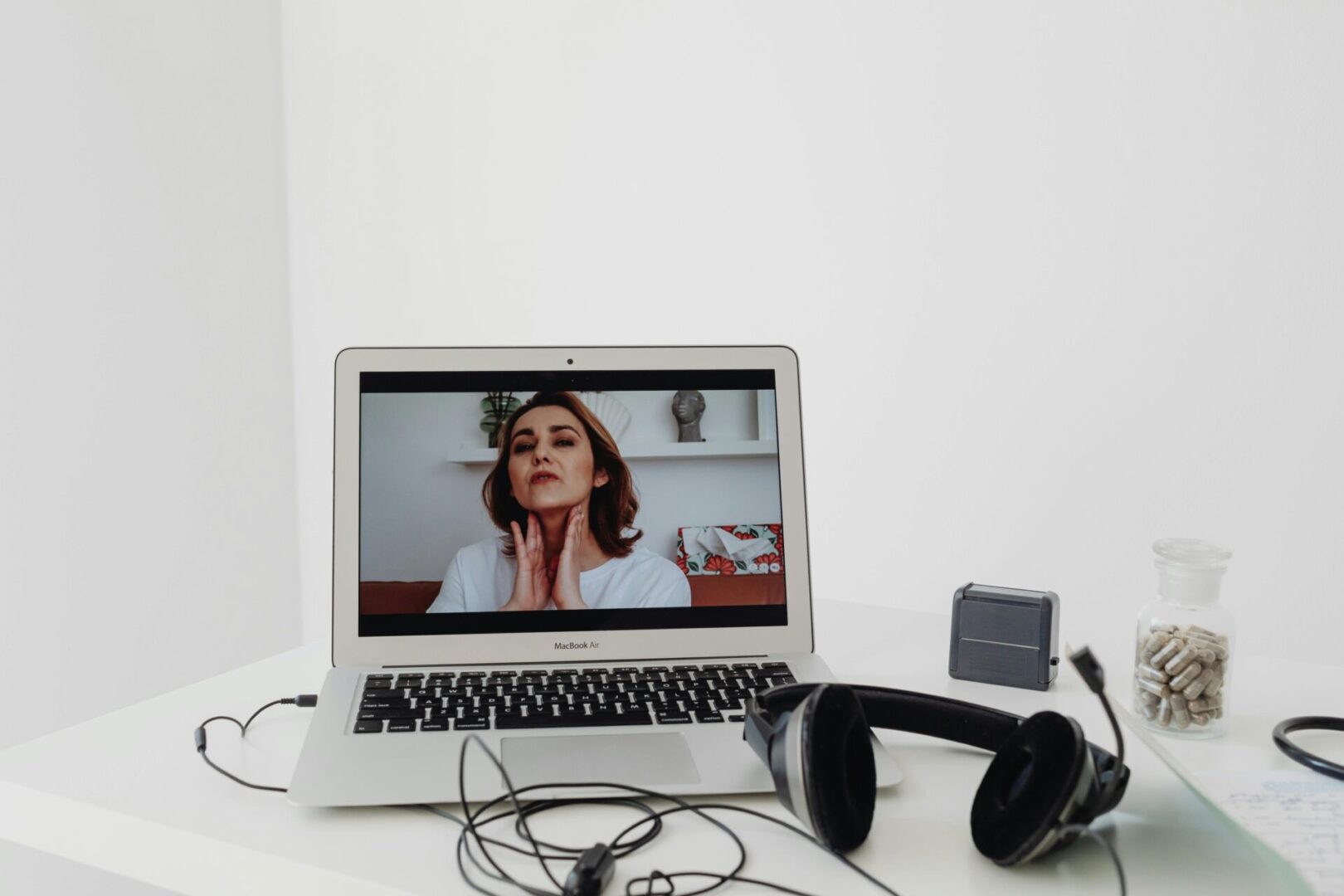 A laptop displays a woman speaking during a video call. Headphones, a small speaker, and a jar of capsules are on the white desk.