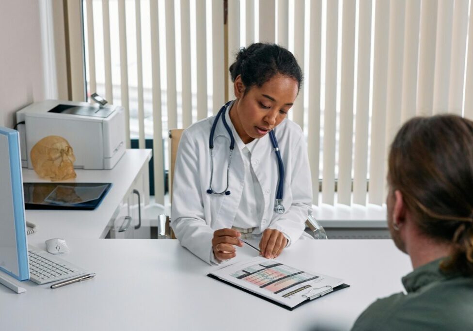 A doctor in a white coat reviews a chart and discusses information with a patient sitting across a desk in a medical office.