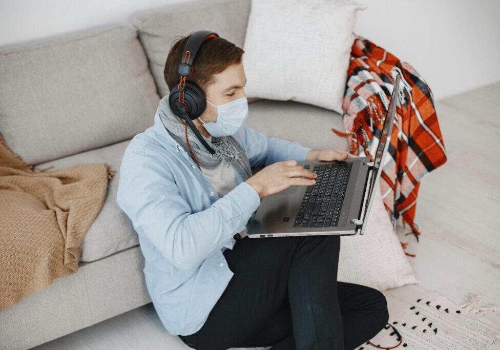 A man wearing a face mask and headphones sits on the floor by a couch, using a laptop.