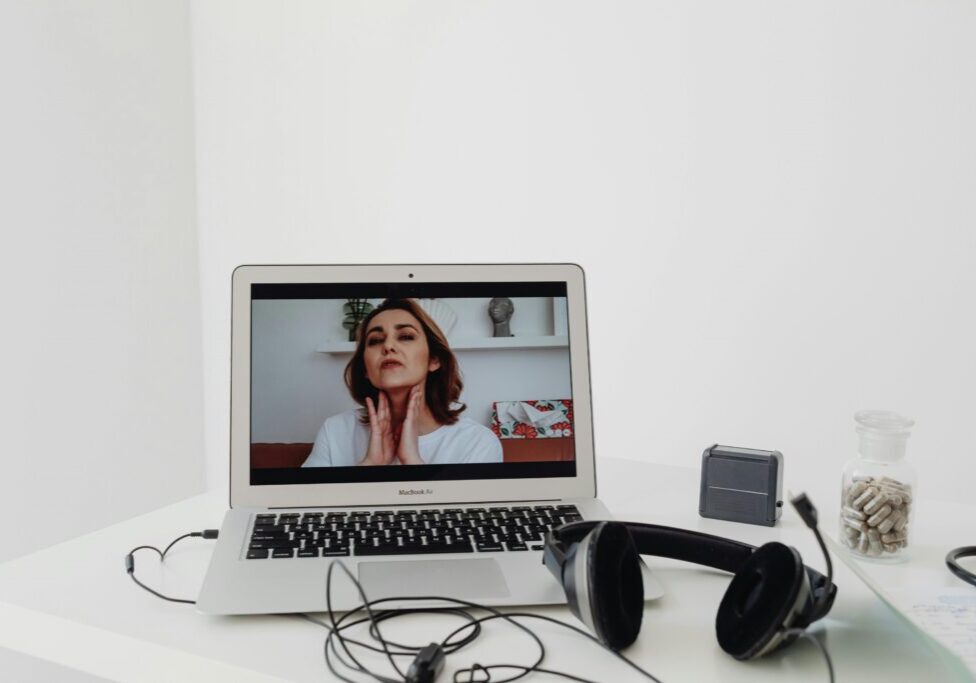 A laptop displays a woman speaking during a video call. Headphones, a small speaker, and a jar of capsules are on the white desk.